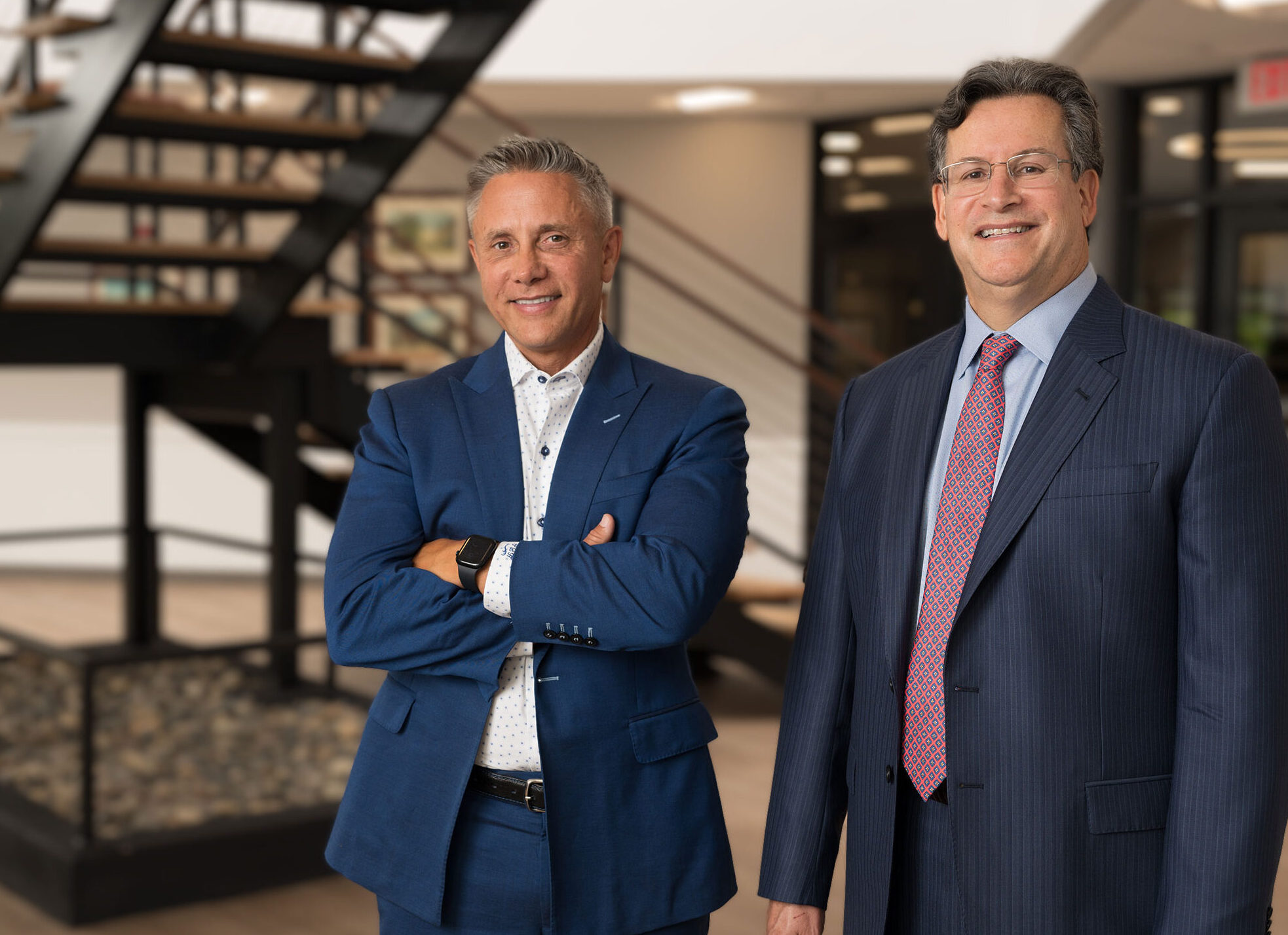 Harding and Mazzotti stand facing the camera in an office building, a set of iron stairs can be seen behind them.